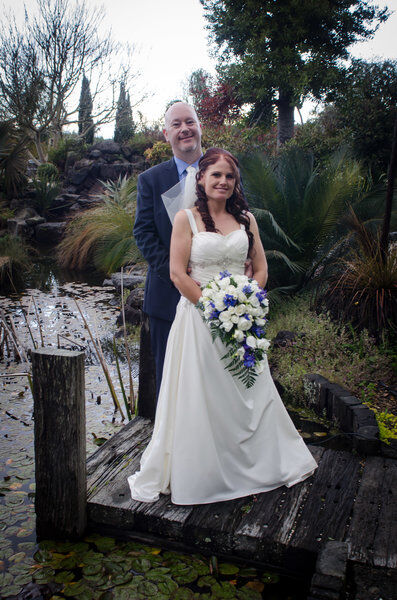 Bride and groom standing on jetty beside lake with bride holding bouquet